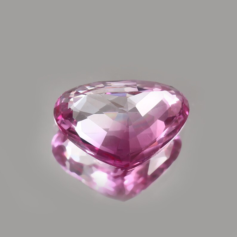 May include: A heart-shaped, faceted pink gemstone with a reflective surface. The gem is a vibrant shade of pink, with multiple facets catching the light. The gem is displayed on a reflective surface, creating a mirrored effect.