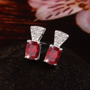 Puede incluir: Pendientes de plata con piedras preciosas rectangulares de color rojo intenso. Cada pendiente presenta una sección rectangular más pequeña sobre la piedra roja, incrustada con pequeños cristales transparentes. Los pendientes se exhiben sobre una superficie oscura y texturizada.