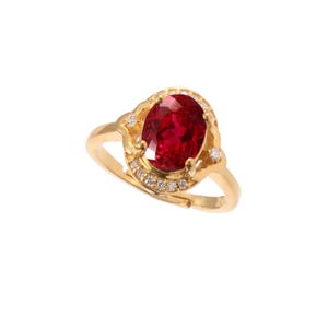 Può includere: Un anello d'oro con una grande gemma rossa brillante a taglio ovale. La gemma è circondata da pietre più piccole e trasparenti e incastonata in una fascia d'oro ornata. L'anello è presentato su sfondo bianco.