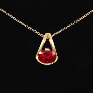 Könnte beinhalten: Eine Goldhalskette mit Anhänger, bestehend aus einem leuchtend roten Edelstein im Ovalschliff, der in einen dreieckigen Goldrahmen gefasst ist. Der Anhänger hängt an einer feinen Goldkette und zeigt eine polierte Oberfläche sowie facettierte Kanten.