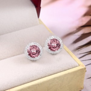 Puede incluir: Un par de pendientes de botón con grandes piedras rosas redondas rodeadas por un halo de piedras claras más pequeñas. Los pendientes se exhiben en una caja de joyería beige con interior rojo.