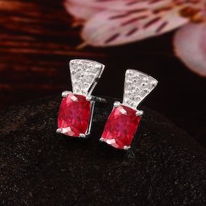 May include: A pair of silver stud earrings. Each earring features a rectangular, bright red gemstone, with a fan-shaped top section set with small, clear stones. The earrings are displayed against a dark, textured surface.