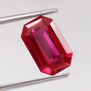 May include: A vibrant red emerald-cut gemstone, held in tweezers, with a polished, faceted surface.