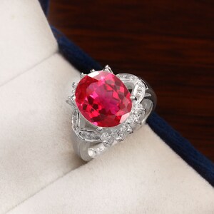 May include: A silver ring featuring a large oval red gemstone, surrounded by smaller clear gemstones.