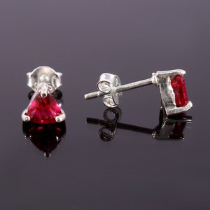 Puede incluir: Un par de pendientes de plata con gemas rojas vibrantes. Un pendiente presenta una piedra triangular, mientras que el otro tiene una piedra rectangular. Los pendientes están sobre una superficie negra reflectante, resaltando su brillo.