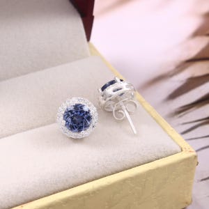 May include: A pair of silver stud earrings with a round, blue gemstone center, surrounded by a halo of smaller, clear stones. The earrings are displayed in a beige jewelry box with a gold trim.