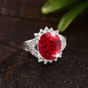 Pode incluir: Um anel de prata com uma grande pedra de rubi ovalada cercada por um halo de pedras menores e transparentes.