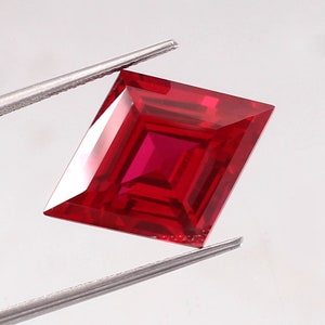 May include: A deep red square-shaped gemstone with a faceted cut. The gemstone is held by tweezers.