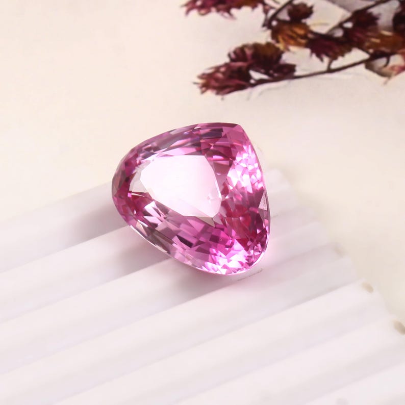 May include: A faceted, heart-shaped pink gemstone. The gem is a vibrant shade of pink, with multiple facets that reflect light. The gemstone is set against a white background, with a few dried flowers in the background.