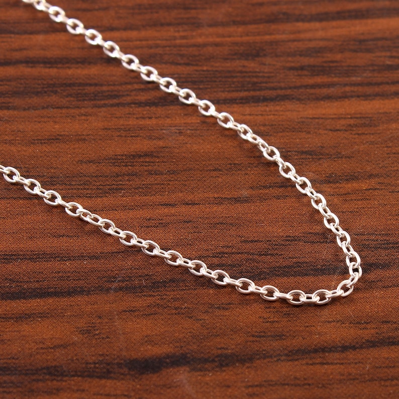 Puede incluir: Un collar de cadena de plata con un dise&ntilde;o delicado de eslabones redondeados. La cadena est&aacute; sobre una superficie de madera marr&oacute;n.