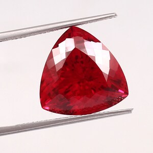 May include: A triangular, deep red gemstone is held by silver tweezers against a white backdrop. The faceted surface of the gem reflects light, highlighting its multiple facets and rich colour.