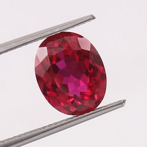 May include: A vibrant red oval gemstone with a faceted cut, held by a pair of tweezers.