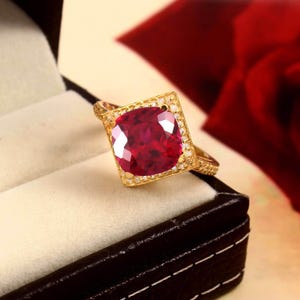 Può includere: Un anello d'oro con una grande pietra preziosa rosso scuro a taglio quadrato. La pietra preziosa è circondata da pietre più piccole e chiare e incastonata in una fascia d'oro dettagliata. L'anello è esposto in un cofanetto nero.
