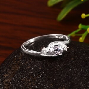 May include: A silver ring featuring a central oval-shaped, pale stone, with smaller round stones either side. The band has a flowing, elegant design. The ring is displayed on a dark, textured surface.