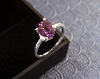Può includere: Un anello d'argento con una grande pietra preziosa viola a taglio ovale. L'anello ha un design classico, con la gemma tenuta in posizione da quattro griffe. È presentato in un cofanetto nero, su uno sfondo blu a motivi.