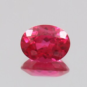 Puede incluir: Una gema de rubí facetada de forma ovalada. La gema es de un color rojo vibrante y saturado con un alto grado de claridad. El rubí se encuentra sobre un fondo claro, con su reflejo visible debajo.