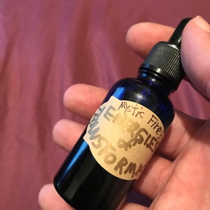 May include: A dark blue glass bottle with a black dropper top. The bottle has a label with the words "Mystic Fire's Energies of Transformation" in handwritten style. The bottle is held in a hand.