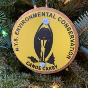Puede incluir: Adorno redondo y amarillo con el texto "N.Y.S. ENVIRONMENTAL CONSERVATION CANOE CARRY" en negro. El adorno presenta una silueta negra de una persona que lleva un kayak. El adorno está colgado de un árbol de Navidad.