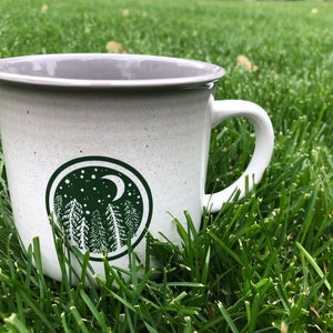 May include: White ceramic mug with a green design of a crescent moon and pine trees in a circle. The mug is sitting on green grass.