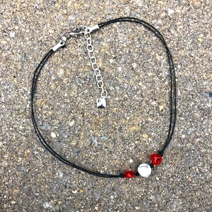 May include: A black cord necklace with a silver heart charm and three beads: two red and one white.