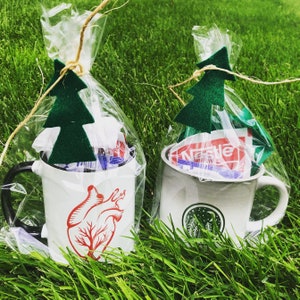 May include: Two white ceramic mugs with different designs. One mug has a red heart with a tree inside and the other has a forest scene with a moon. Both mugs are wrapped in clear plastic with green paper tree ornaments.