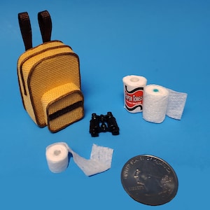 May include: Miniature yellow and brown backpack, black binoculars, rolls of paper towels, and a quarter on a blue background. The paper towels have the words "Paper Towels" printed on them.