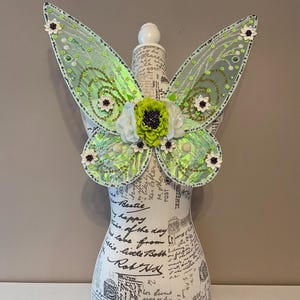 May include: Iridescent green fairy wings with a floral design, displayed on a mannequin. The wings feature a gradient of green and white, with small white and purple flowers. The wings are decorated with sequins and a central flower arrangement.