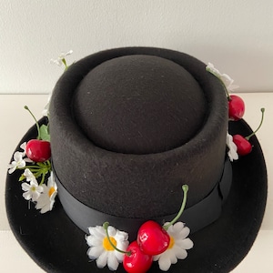 Può includere: Un cappello fedora in feltro nero con un nastro nero e decorazioni di margherite bianche e ciliegie rosse.