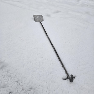 May include: A long, metal spatula with a perforated head lies on a snowy surface. The spatula has a twisted handle and a hook at the end.