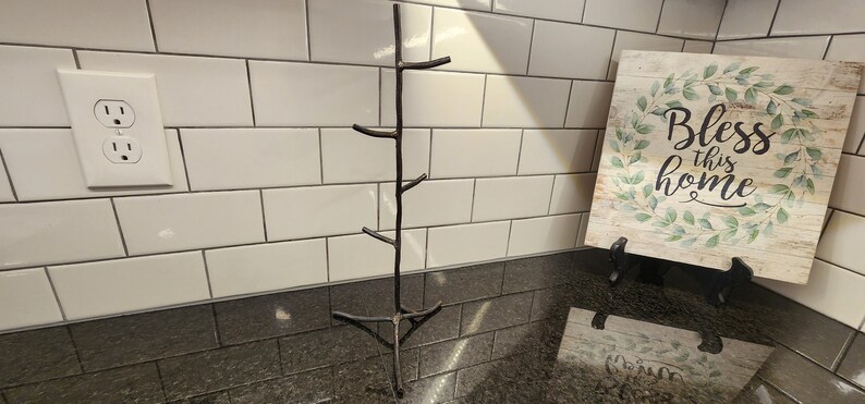 A black metal twig-shaped stand with five branches. The stand is on a black countertop with white subway tile behind it. A sign with the words 'Bless this home' is on the counter to the right of the stand.