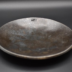 May include: A small, round, metal dish with a weathered, mottled surface. The dish has a slightly raised edge and a dark, aged appearance. The text "LHT I" is stamped on the rim. The dish is set against a dark background.