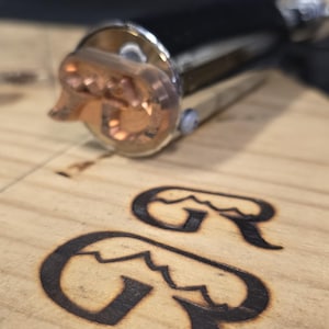 May include: A metal branding iron with a copper head featuring a mountain-shaped letter G design. The tool is positioned above a light-colored wooden surface that displays two clear, dark-burned impressions of the same mountain-shaped letter G logo.