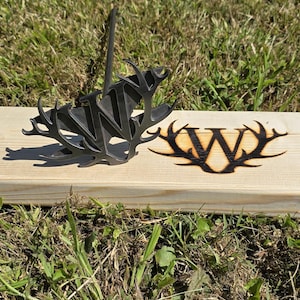 May include: A metal branding iron with the letter "W" and antler design, along with a branded wooden block with the same design. The branding iron is black and sits on a light-colored wooden plank.