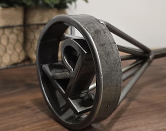 May include: A metal branding iron with the letters "RV" in a circular design. The iron is made of dark, weathered metal and sits on a wooden surface. The background includes greenery.