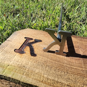May include: A metal branding iron shaped like the letter "K" on a wooden surface. The letter "K" is also burned into the wood.