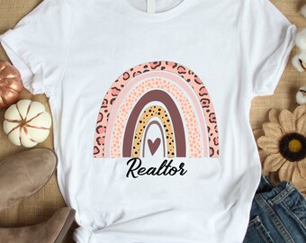 May include: White t-shirt featuring a colorful rainbow design with leopard print accents. The word "Realtor" is printed in black script below the rainbow. The shirt is displayed with jeans and boots.
