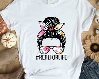 May include: White t-shirt with a floral print design of a woman with a messy bun, sunglasses, and a headband. The text "#REALTORLIFE" is printed below the design.