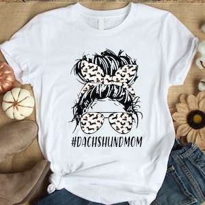 Pu&ograve; includere: Maglietta bianca con un'illustrazione in bianco e nero di una donna con un chignon disordinato e occhiali da sole. Il chignon e gli occhiali da sole sono decorati con un motivo di cani bassotti. Il testo "#dachshundmom" &egrave; stampato sotto l'illustrazione.