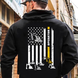 Könnte beinhalten: Schwarzer Hoodie mit einem weißen und gelben amerikanischen Flaggen-Design. Die Flagge hat einen gelben Streifen auf der rechten Seite mit dem vertikal gedruckten Wort "OPERATOR". Die Flagge zeigt mehrere Bilder von Baumaschinen, darunter Bagger.