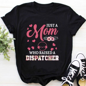Könnte beinhalten: Schwarzes T-Shirt mit rosa Schriftzug "Just a Mom who raised a Dispatcher". Der Text ist von rosa Herzen und Blumenmustern umgeben.
