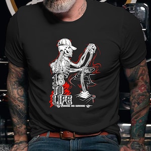 Op de afbeelding: Zwart T-shirt met een witte skeletillustratie die een auto bestuurt. De tekst "18 TO LIFE" is in rood en wit gedrukt, met daaronder de woorden "DRIVING AND SURVIVING".