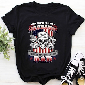 Op de afbeelding: Zwart T-shirt met een afbeelding van een schedel die een baseballpet draagt en sleutels vasthoudt. De schedel bevindt zich voor een Amerikaanse vlag. De tekst "Some people call me a Mechanic. The most important call me Dad." staat op het T-shirt gedrukt.