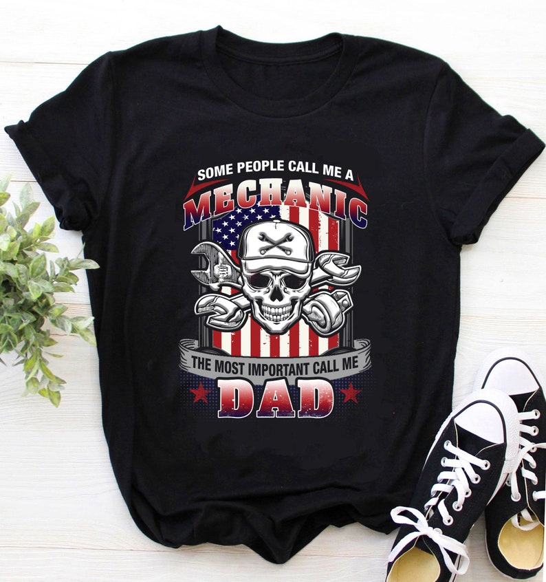 Op de afbeelding: Zwart T-shirt met een afbeelding van een schedel die een baseballpet draagt en sleutels vasthoudt. De schedel bevindt zich voor een Amerikaanse vlag. De tekst "Some people call me a Mechanic. The most important call me Dad." staat op het T-shirt gedrukt.