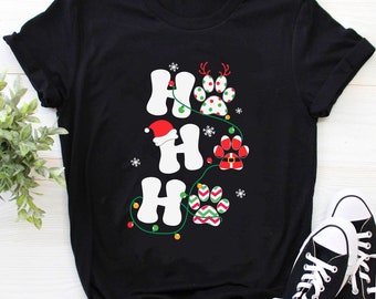 Könnte beinhalten: Schwarzes T-Shirt mit dem Text "HO HO HO" in Weiß, wobei die Buchstaben O durch Pfotenabdrücke ersetzt werden. Die Pfotenabdrücke sind mit Weihnachtsbeleuchtung, einem Weihnachtsmannhut und Rentiergeweih verziert. Ein Paar schwarze Turnschuhe mit weißen Schnürsenkeln im Vordergrund.
