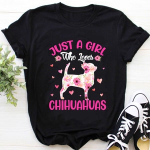 May include: Black t-shirt with the text "JUST A GIRL Who Loves CHIHUAHUAS" in pink. The shirt features a floral Chihuahua design with pink flowers and hearts. A pair of black and white sneakers are in the bottom right corner.