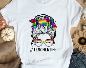 May include: White t-shirt with a rainbow heart and a woman's face with glasses. The text "#TEACHERLIFE" is printed below the image.