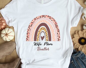 May include: White t-shirt with a pink, brown, and yellow rainbow design. The rainbow has a heart in the center and is surrounded by leopard print. The text "Wife Mom Realtor" is below the rainbow.