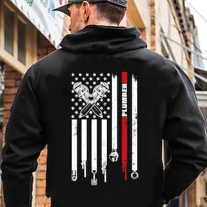 May include: Black hoodie with a white and red American flag design. The flag has crossed wrenches and the word "PLUMBER" printed vertically on the right side.