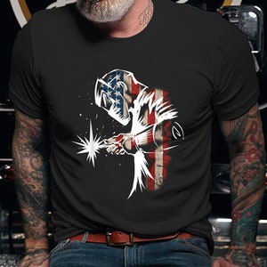May include: A black t-shirt with a white silhouette of a welder wearing a welding mask. The welder is standing in front of an American flag.