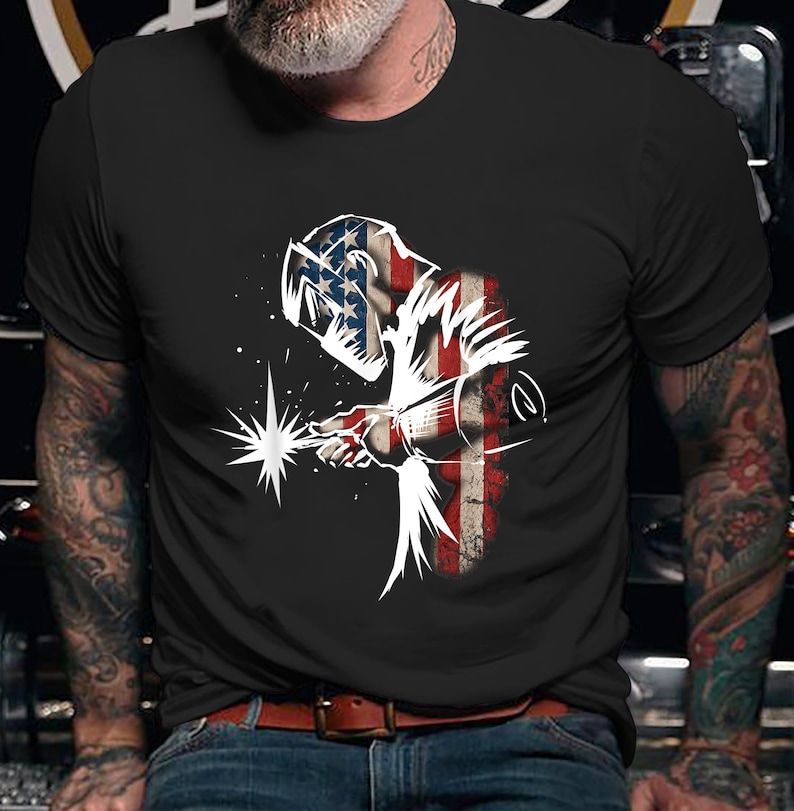 May include: A black t-shirt with a white silhouette of a welder wearing a welding mask. The welder is standing in front of an American flag.